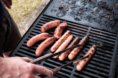 Genç beyaz adam elleriyle ızgarada sosis hazırlıyor.