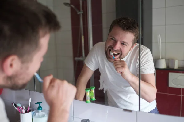 Young man with toothbrush cleaning teeth and looking to mirror at home bathroom