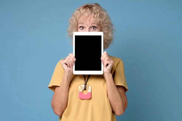 Зріла жінка ховає обличчя за портативним комп'ютерним пристроєм на синьому фоні — стокове фото