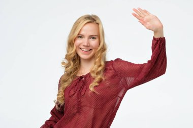 Genç sarışın kadın mutlu bir şekilde gülümsüyor, merhaba ya da güle güle diyor.