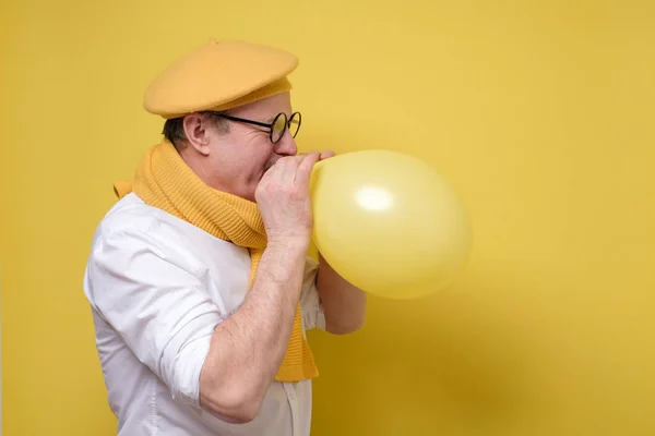 Man in yellow beret and scarf getting ready to the party blowing a balloon. — Stockfoto