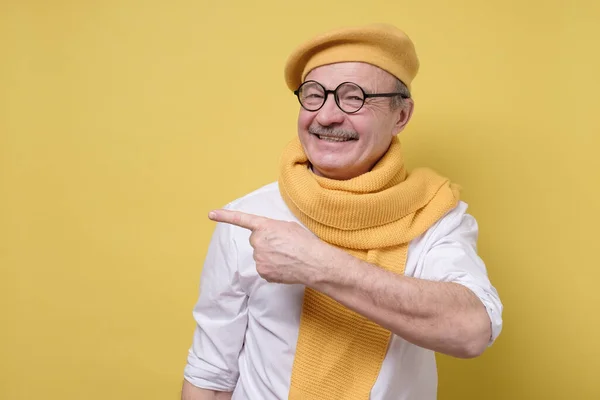 Självsäker senior leende man i gul basker och halsduk peka bort. — Stockfoto