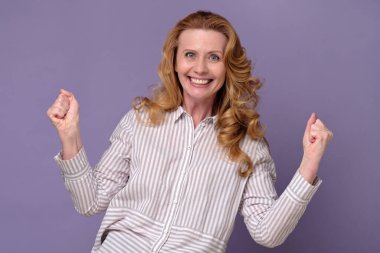 Heyecanlı ve mutlu bir başarı. Yumrukları havada genç bir kadın, başarısını kutluyor. Mor arkaplanda stüdyo çekimi.