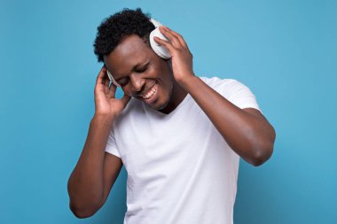 Beyaz kulaklıklı ve tişörtlü müzik dinleyen genç bir Afro-Amerikan. Stüdyo resmi mavi duvarda.