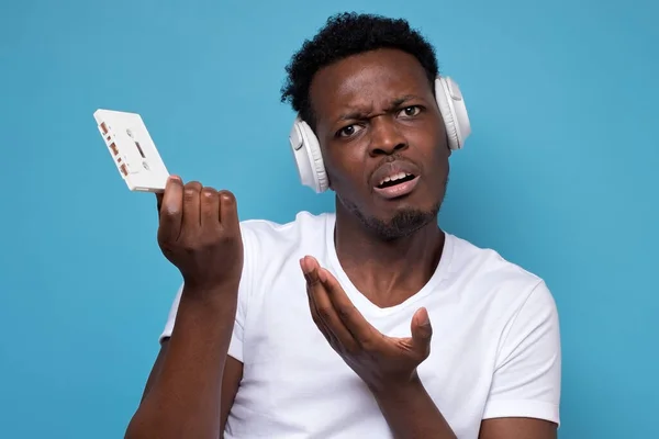 Young confused millenial african man with white headphones holding retro cassette asking how to use it on blue background. Old retro technology concept. Studio shot