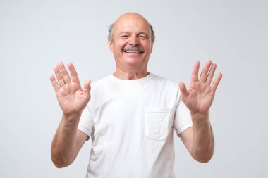 Heyecanlı kel adam beyaz bir arka planda neşeli bir şekilde izole edilmiş on parmağını ve gülüşünü gösteriyor, ellerini kaldırıp onu güldürmeyi kesmesini diliyor. Stüdyo çekimi