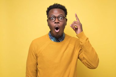 Sarı arka planda gözlüklü genç Afrikalı bir adam yukarı doğru fikir ya da eureka hareketi gösteriyor. Öğrenci ilginç bir plan yapıyor. Stüdyo çekimi