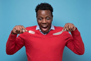 Çatal ve bıçak tutan kırmızı kazaklı çılgın Afro-Amerikan adam..