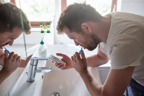 Νεαρός καυκάσιος άνδρας στο μπάνιο βουρτσίζει τα δόντια του ενώ στέλνει μηνύματα στο κινητό τηλέφωνο — Φωτογραφία Αρχείου