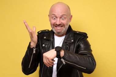 Mature italian man in black leather jacket impatiently pointing to his watch. You are late concept. Isolated on yellow wall clipart