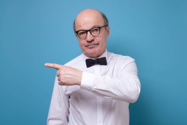 Kendinden emin kıdemli adam işaret etti. Ciddi, olgun bir erkek, mavi stüdyo arka planında parmağını bir kenara koyuyor.