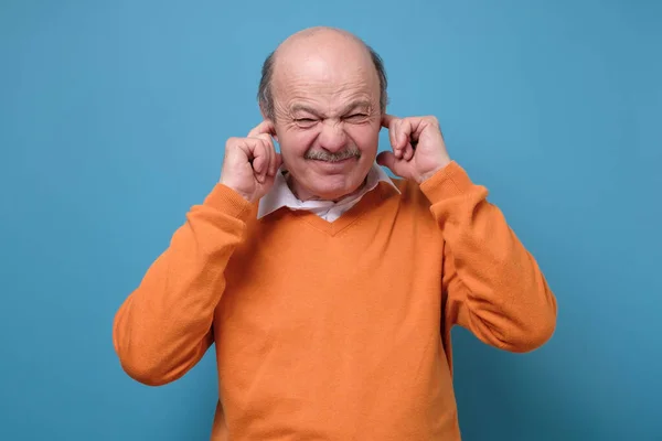 Stop that loud noise concept. Mature man suffering from loud music and pain trying to plug ears against blue background. Sadness emotion concept