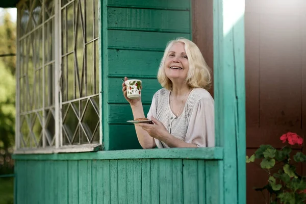 晴れた日に家の入り口とお茶を飲むカップの近くに立っている上のシニア白人女性の肖像画 — ストック写真
