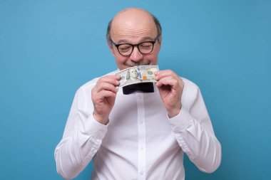 Gözlüklü kıdemli beyaz adam bir dolar banknot tutuyor ve mavi arka planda onu kokluyor. İş adamının nakit parası var. Zenginlik ve refah kavramı. Kolay ve çabuk para kazan..