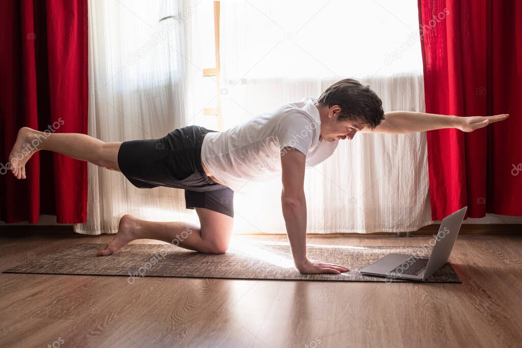 Young caucasian man practices yoga asana chakravakasana or bird pose at the living room at home using laptop for online lesson.