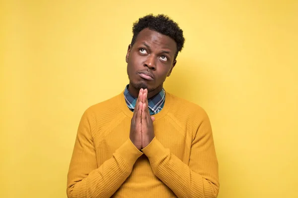 Please forgive me. Praying young Afro American male pressing hands together, having guilty look, asking for forgiveness, hoping for better on yellow wall. Negative human emotion.