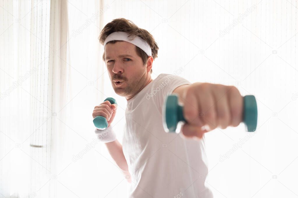 Funny mature man exercising at home holding weights near window at living room.