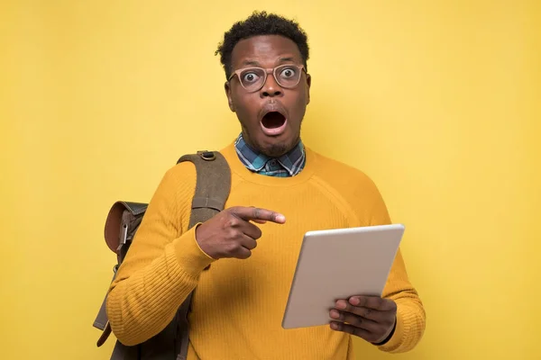 Happy african american college student in glasses holding tablet on yellow wall.