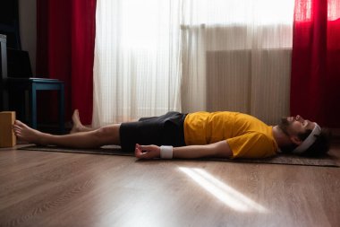 Genç adam yerde meditasyon yapıyor ve oturma odasında Shavasana pozu veriyor..