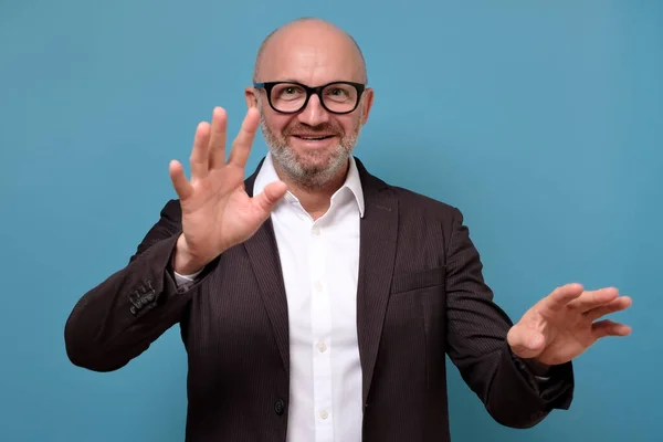 Handsome mature guy in glasses and suit reaching out to the camera isolated. Give it to me. Studio shot on blue wall.