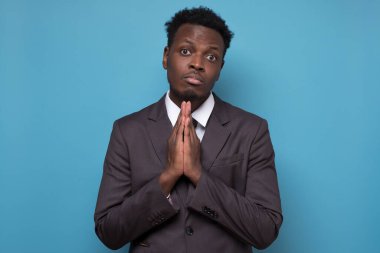 Endişeli Afro-Amerikan takım elbiseli genç adam ellerini birleştirip mavi stüdyo arka planında izole edilmiş dua için af dile.