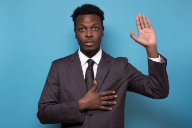 Gerçeği söyleyeceğine yemin et. Samimi Afro-Amerikan erkek takım elbiseli, avuç içini açıp göğsüne dokunuyor, bana dürüst olacağına, mavi arka planda duracağına söz veriyor.