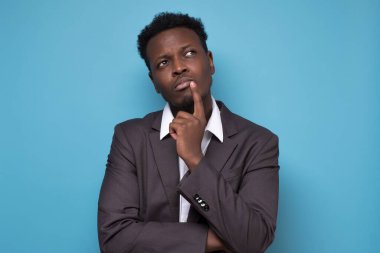 Takım elbiseli genç Afrikalı adam mavi arka planı düşünüyor ve yeni bir plan yapıyor.
