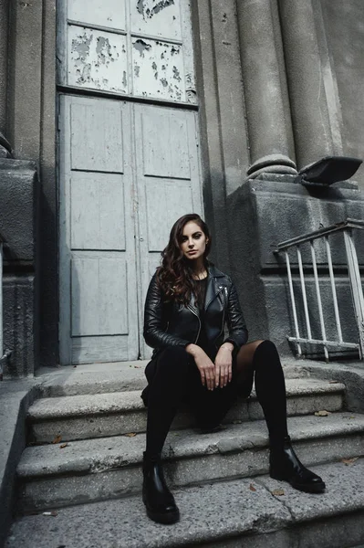 Eastern beautiful woman wearing biker jacket poses in backyard of vintage apartment house — Stock Photo, Image