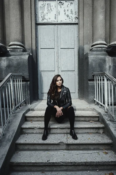 Eastern beautiful woman wearing biker jacket poses in backyard of vintage apartment house — Stock Photo, Image