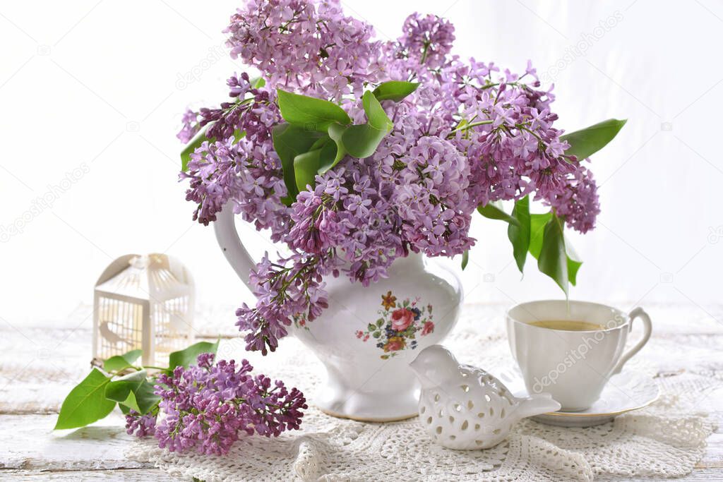 purple lilac blossom branches in porcelain vase and cup of coffee on white table in vintage style