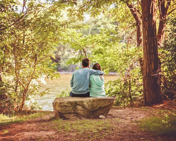 一对相爱的情侣坐在河岸上 男人在温暖的夏日把手臂披在女人的身上 — 图库照片