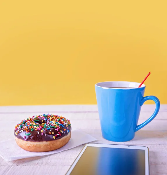 Chocolate doughnut with colorful sprinkles on a napkin next to a blue coffee cup and a tablet on a wooden table with a yellow background — Stock Photo, Image
