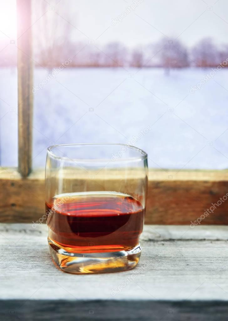 glass of whiskey or other alcohol in front of a cabin window on a cold winter day with trees and mountains in the view 