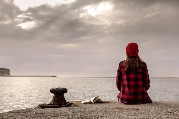 woman with book sitting on pier and enjoying beautiful view of lake at cloudy day 