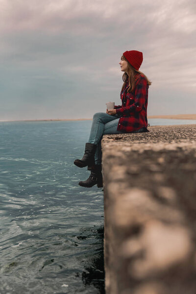 beautiful woman sitting on pier of lake and enjoying day with mug of hot coffee