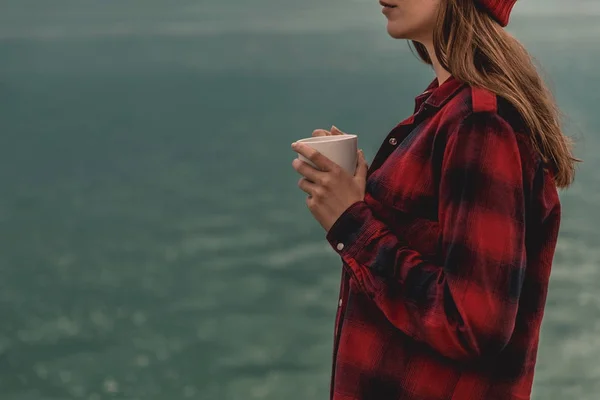 美丽的女人捧着一杯热咖啡站在湖岸边 — 图库照片