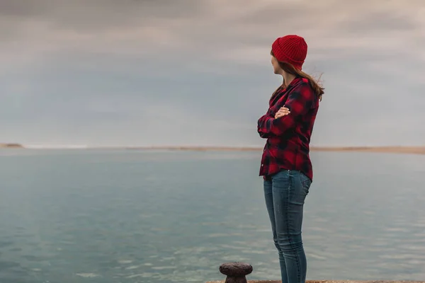 赤い帽子とシャツ寒い曇りの日に湖の桟橋に立っている間で美しい女性 — ストック写真