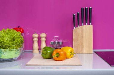 Taze sebzeler doğrama tahtasının üzerinde yatar. Arka planda bir bıçak seti ve biber kapları var..