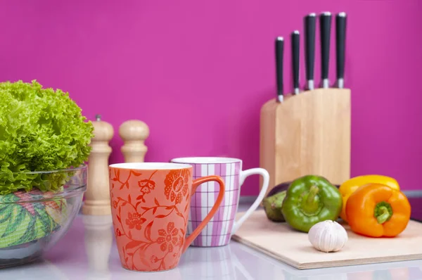 Gri mutfak masasında iki fincan duruyor. Taze sebzeler tahta tahtada yatar. Arka planda bir bıçak seti ve biberlik var.