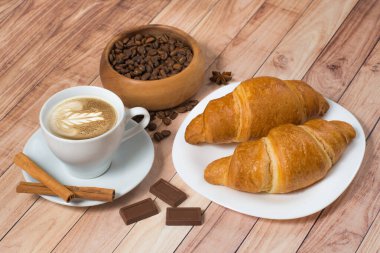 İçinde kruvasan, çikolata, kahve çekirdeği ve ahşap arka planda tarçın olan kahve fincanı. Latte sanatı.