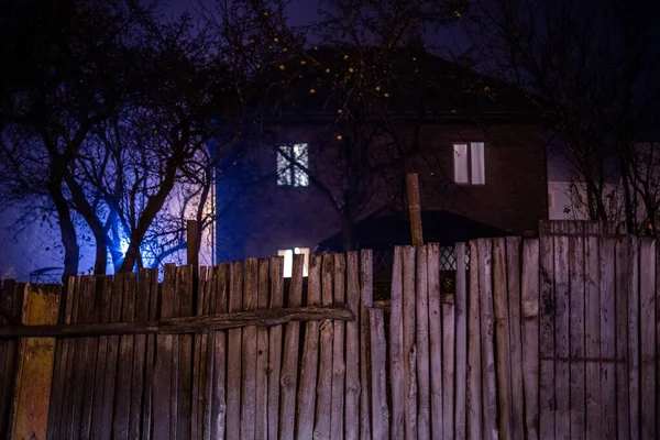Dark foggy night view on wooden fence and spooky house with sele