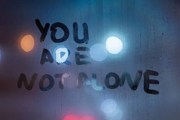 Фраза "ты не один" написана пальцем на ночном мокром стекле с размытыми классическими голубыми огнями на заднем плане — стоковое фото