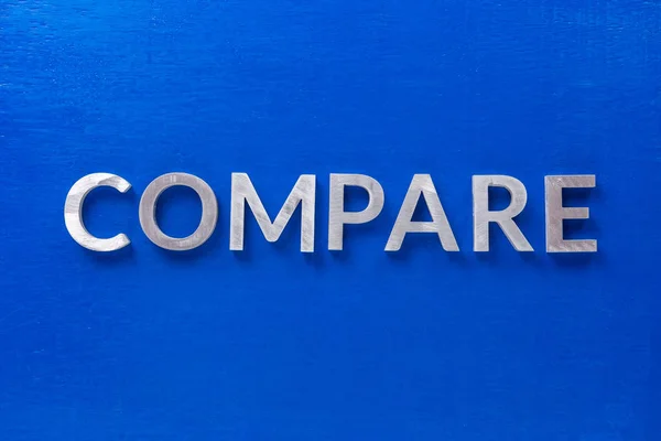 The word compare laid with silver metal characters on blue painted wooden board in flat lay with central composition — 스톡 사진