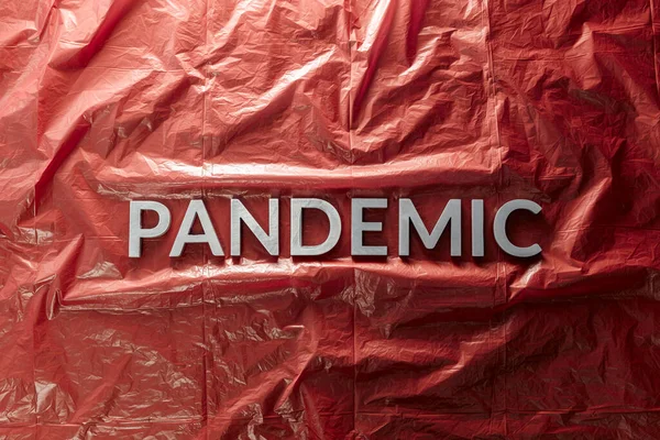 Słowo pandemia ułożone srebrnymi literami na czerwonym pogniecionym tle folii z tworzywa sztucznego w płaskiej kompozycji świeckiej w centrum — Zdjęcie stockowe