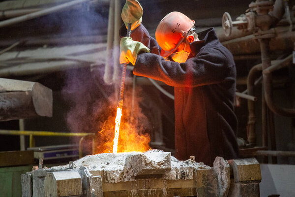 worker with hard hat and face mask stirs liquid metal in a furnace by steel bar - close-up with selective focus and background blur