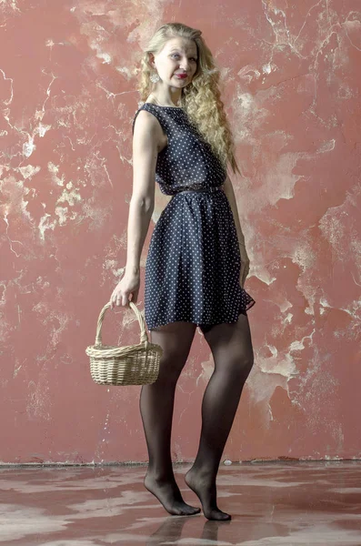 Young girl with blonde curly hair in a long dress with polka dots and stockings dancing with a basket — Stock Photo, Image