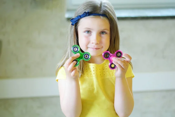 Pequena menina bonita em camiseta amarela está brincando com dois spinners nas mãos Imagens Royalty-Free