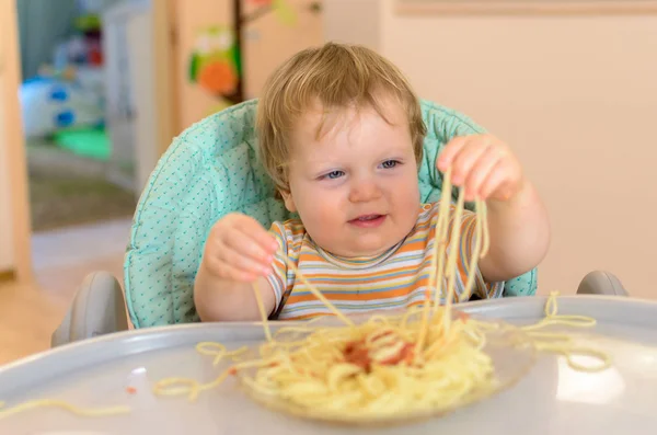 Cute one year old baby eats spaghetti in a children's chair. Royalty Free Stock Images