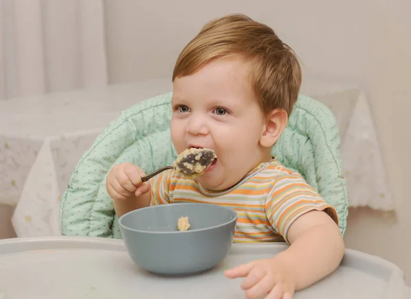 Маленький блондин в полосатой футболке ест кашу ложкой — стоковое фото