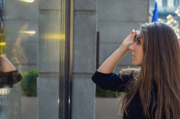 Hermosa chica levanta sus gafas de sol y mira el escaparate o — Foto de Stock